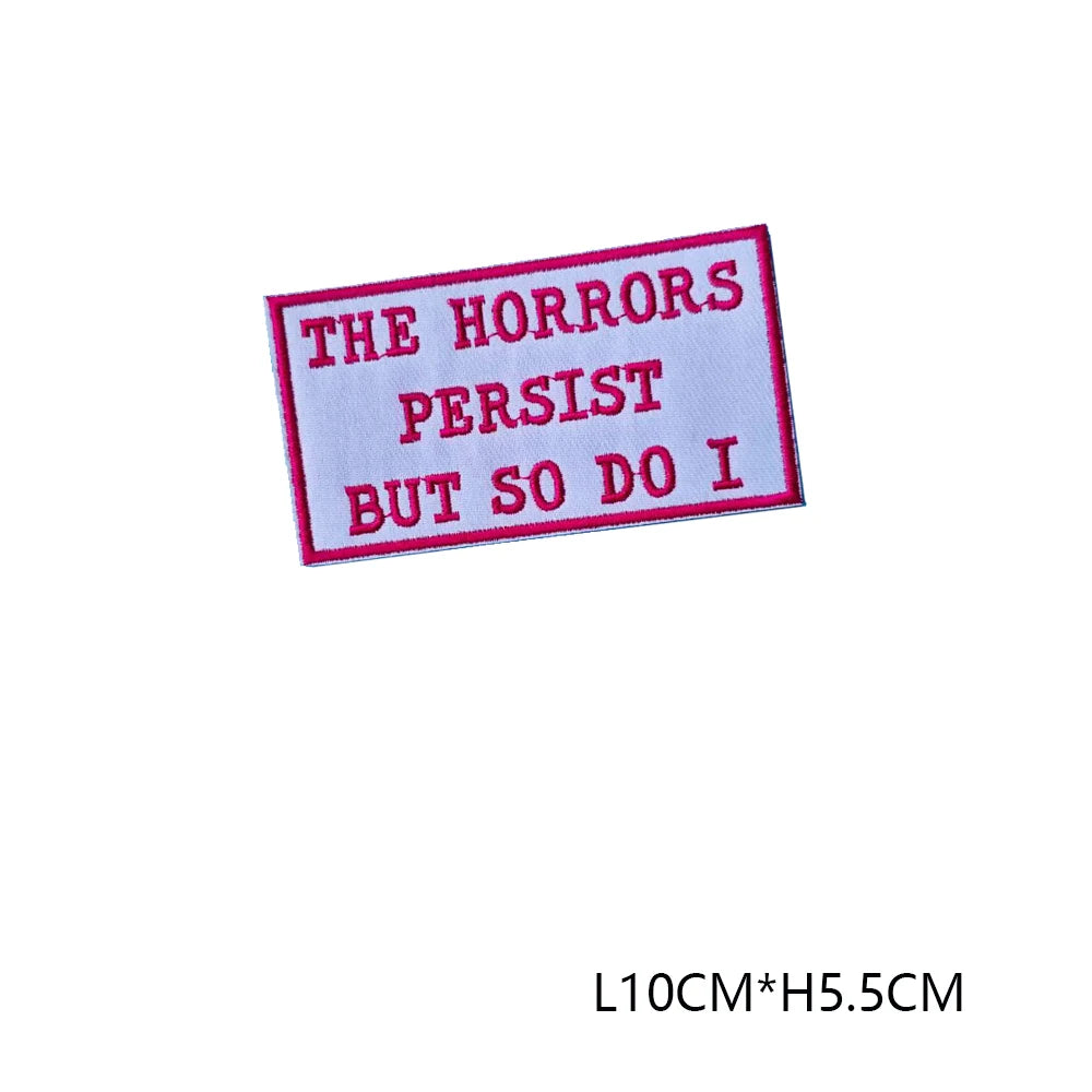 A pink embroidered letter patch with the text THE HORRORS PERSIST BUT SO DO I on a white background.