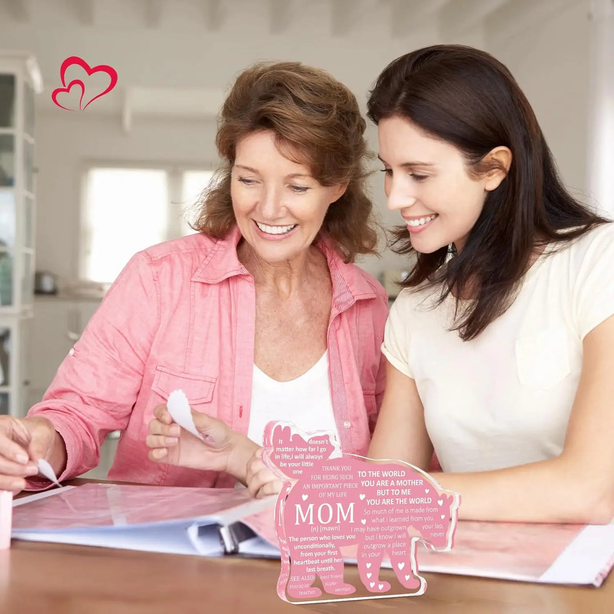 A woman is signing a personalized mom plaque keepsake for a woman in a bright indoor setting.