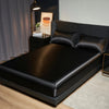 A black satin bedding set is displayed on a bed, next to a dark nightstand and a framed piece of art.