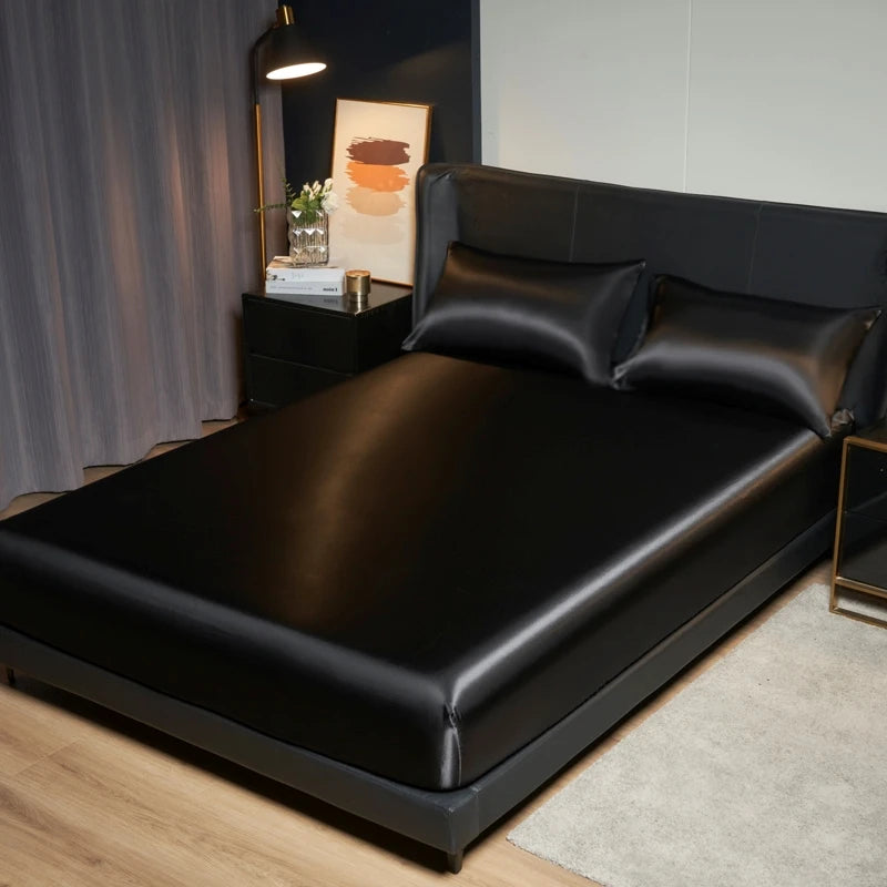 A black satin bedding set is displayed on a bed, next to a dark nightstand and a framed piece of art.