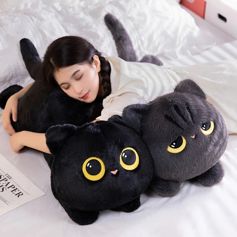 A black cat plush pillow is being held by a person lying in white bedding.