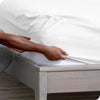 A hand is adjusting a white fitted mattress protector sheet on a wooden bed frame.