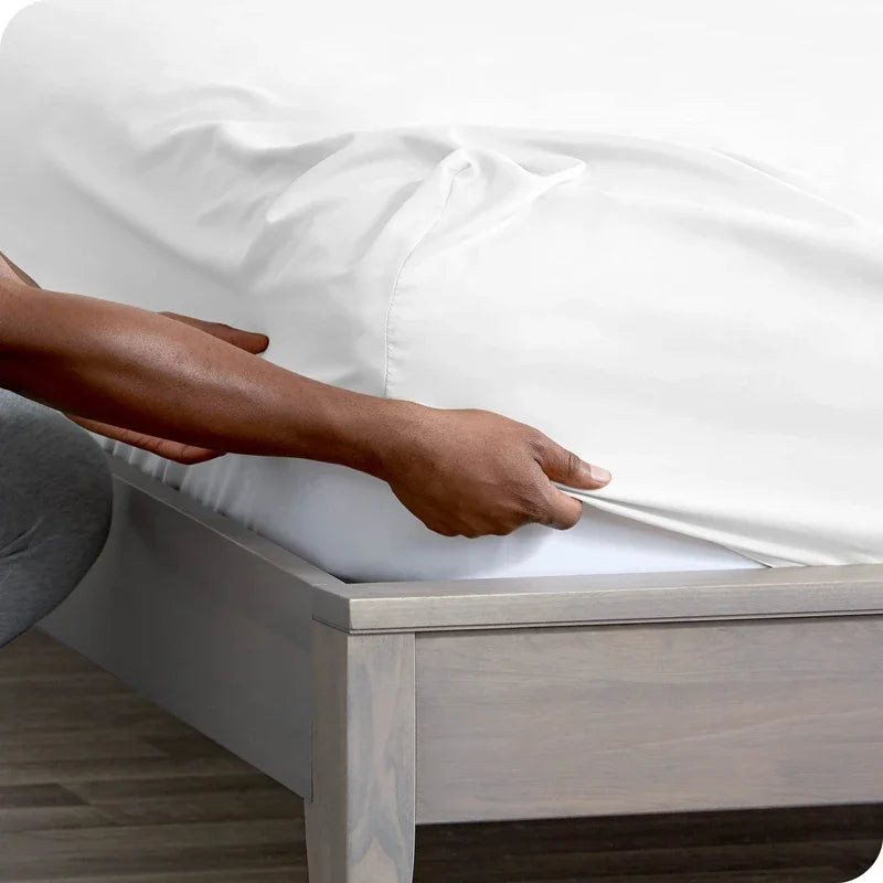 A hand is adjusting a white fitted mattress protector sheet on a wooden bed frame.