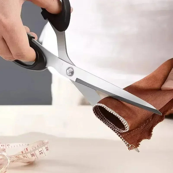 A hand is holding professional heavy duty fabric scissors with silver blades, positioned near a piece of brown fabric and a m