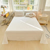 A white organic cotton bedding set featuring a flat sheet, pillowcases, and a stuffed animal on a bed in a bright room.