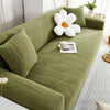 A green stretchable sofa cover with white decorative pillows and a cup on the seat.