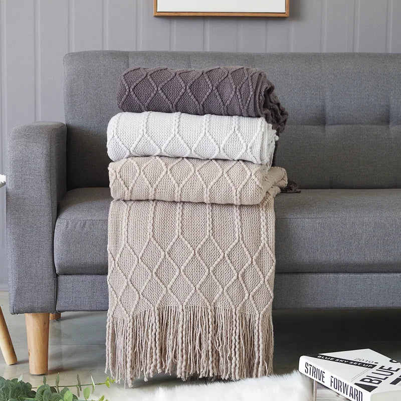 A stack of oversized knitted tassel throw blankets in shades of gray and beige on a sofa.