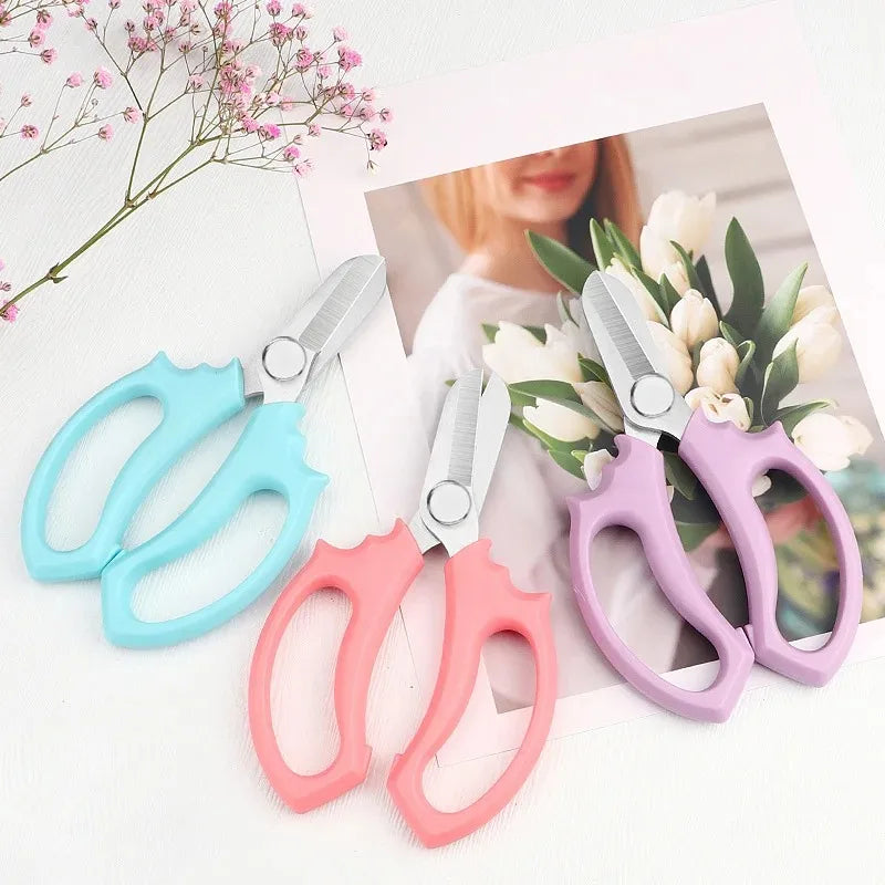 A set of pink and blue precision shears for bonsai and floral work displayed on a white background with a soft floral backdro