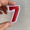 A hand holds a red embroidered number 7 patch with white edges against a light background.