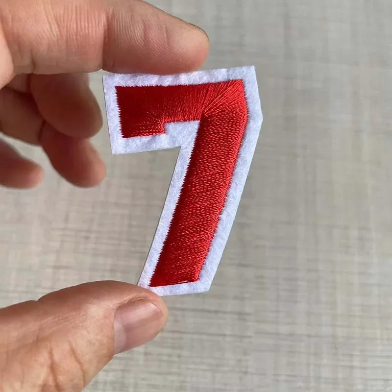 A hand holds a red embroidered number 7 patch with white edges against a light background.