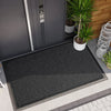 A black non-slip rubber doormat placed on a gray tiled floor near a dark gray door and potted plants.