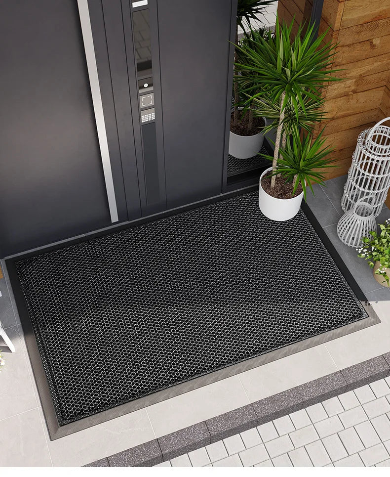 A black non-slip rubber doormat placed on a gray tiled floor near a dark gray door and potted plants.