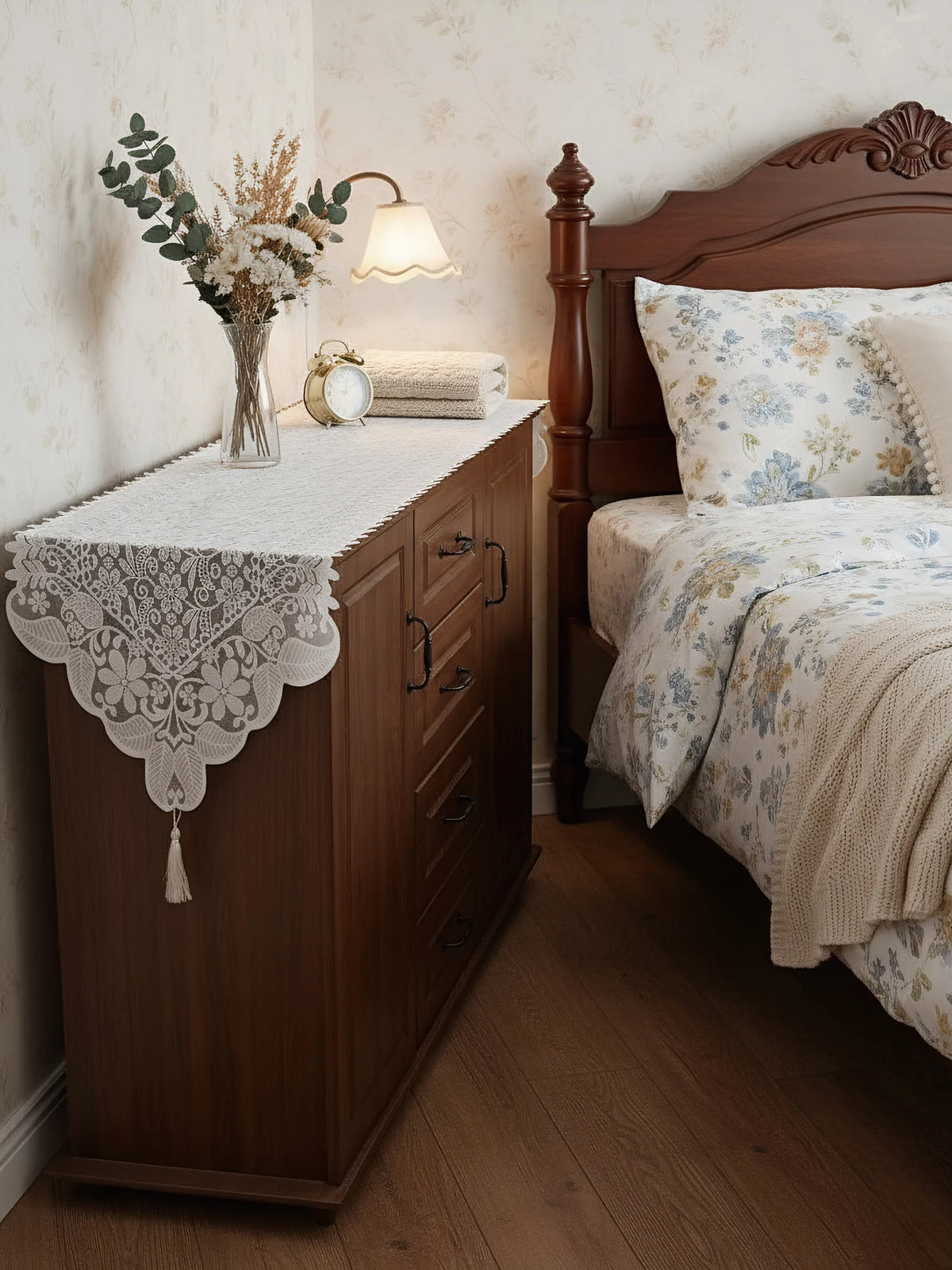A wooden cabinet with a lace table runner and floral arrangement on top, next to a dark wood bed frame.