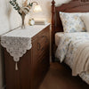 A wooden cabinet with a lace table runner and floral arrangement on top, next to a dark wood bed frame.