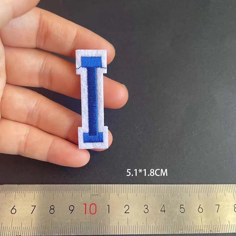 A hand holds a blue and white alphabet embroidery iron-on patch shaped like the letter I, measured against a ruler.