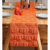 An orange festive Halloween table runner displayed on a wooden table with pumpkins and glassware.