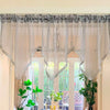A sheer gray beaded curtain with tassels hangs over a window, decorated with lace trim.