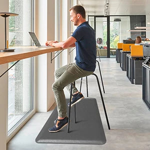 A man sits on an anti-fatigue PVC kitchen mat at a modern desk working on a laptop.