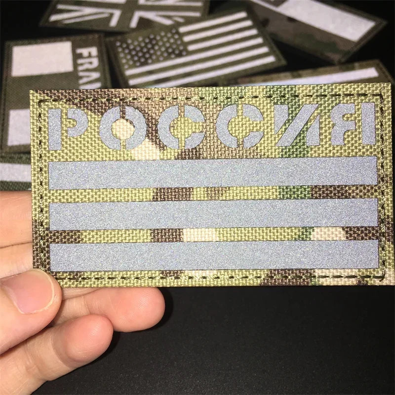 A hand holds a tactical multicam national flag patch with Cyrillic text and horizontal stripes.