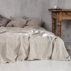 A light beige pure linen flat sheet draped over a bed with soft pillows and a rustic wooden table in a bright room.