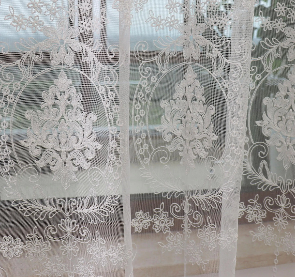 Sheer white embroidered curtains with floral designs covering a glass window.