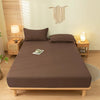 A brown fitted sheet set with matching pillowcases is displayed on a wooden bed frame in a bright room.