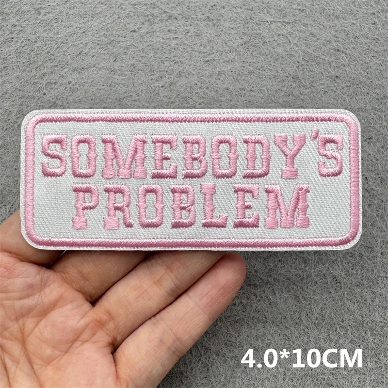 Pink embroidered slogan patch with the text Somebody's Problem on a gray fabric background.