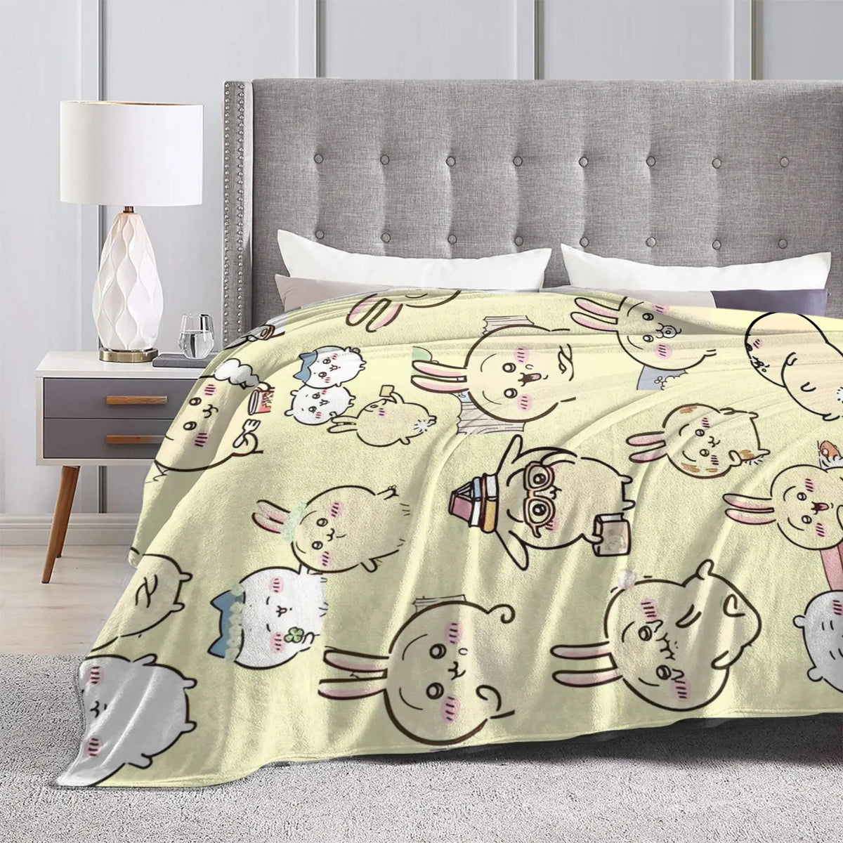 A beige flannel throw blanket featuring a pattern of cute Chiikawa cartoon characters on a bed.