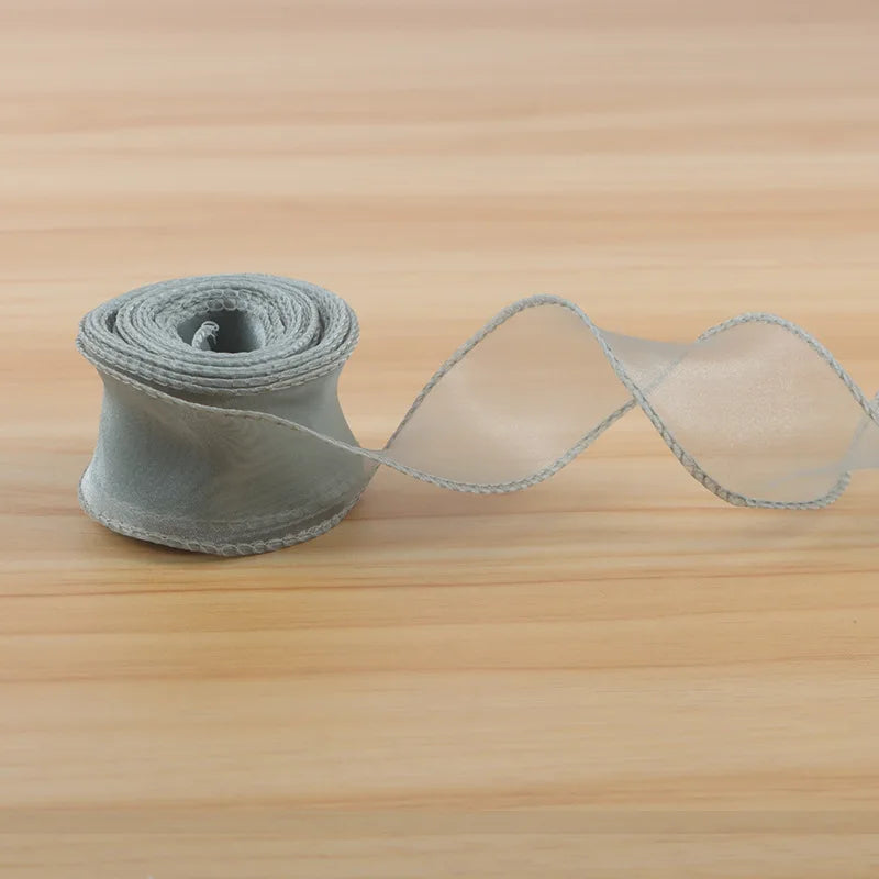 A roll of light gray satin ribbon is unwinding, showing a long, sheer ribbon strip on a wooden surface.