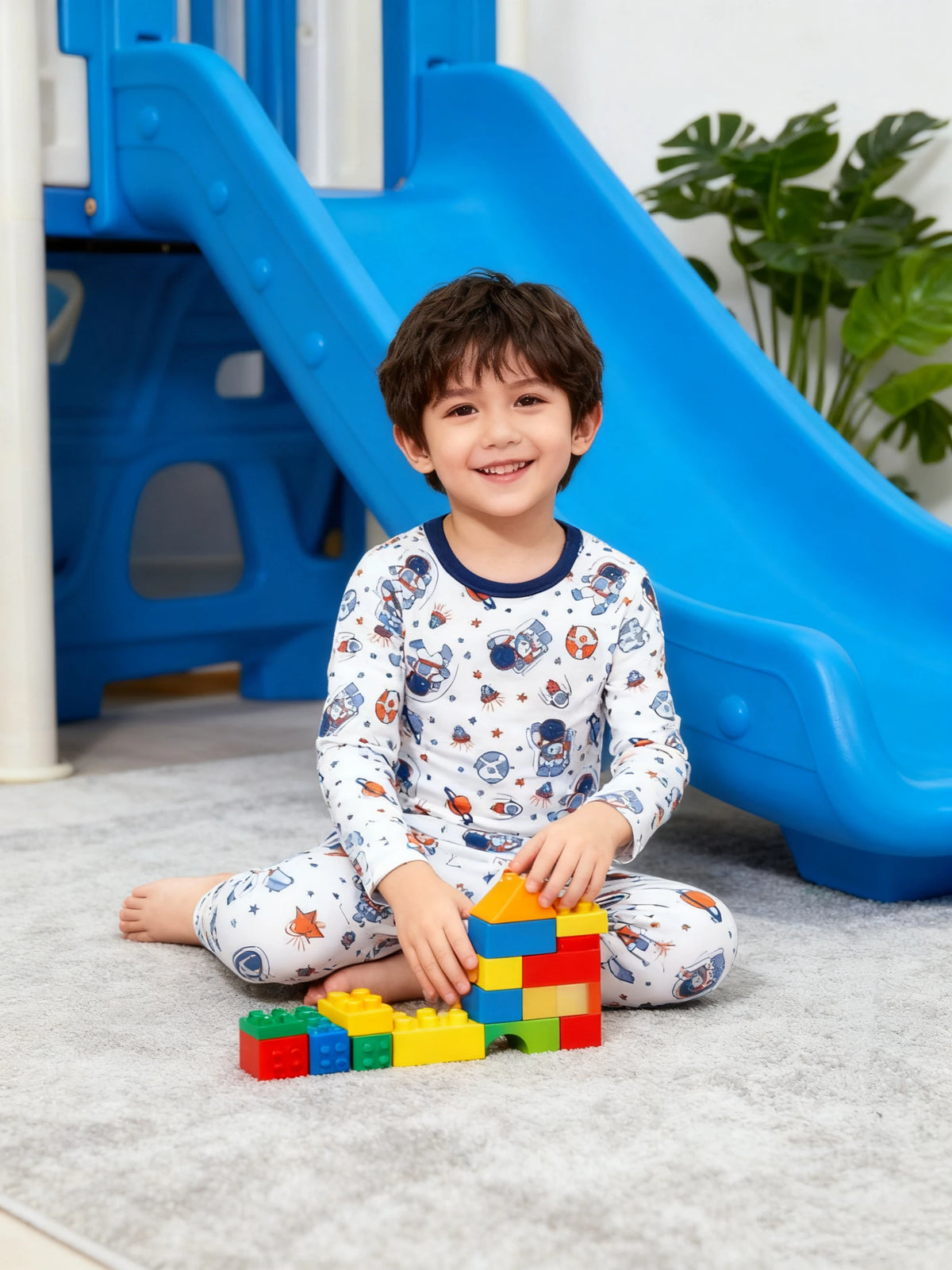 A smiling young boy wearing a white cartoon astronaut pajama set sitting on the floor near a blue slide.