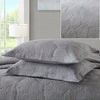 A set of gray embroidered cotton quilted pillow covers on a bed with a white lamp in the background.