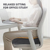 A woman is sitting at a desk using a laptop while resting on an ergonomic U-shaped massage seat cushion.