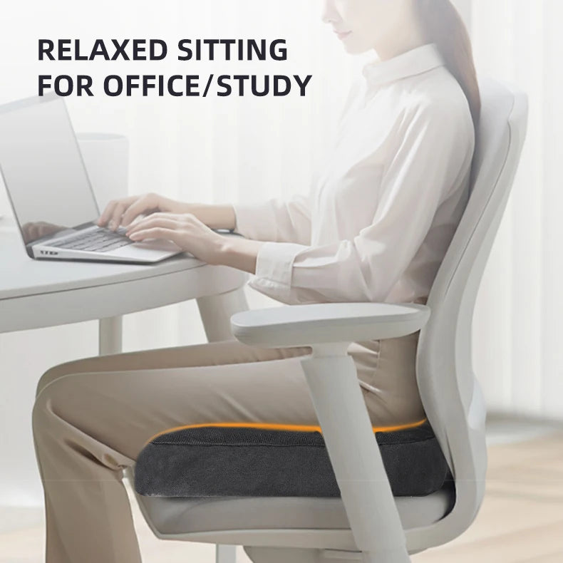 A woman is sitting at a desk using a laptop while resting on an ergonomic U-shaped massage seat cushion.