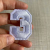A hand holds a white embroidered number 3 patch for apparel.