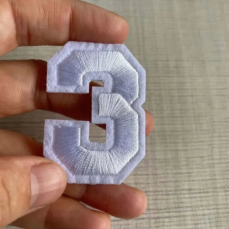 A hand holds a white embroidered number 3 patch for apparel.
