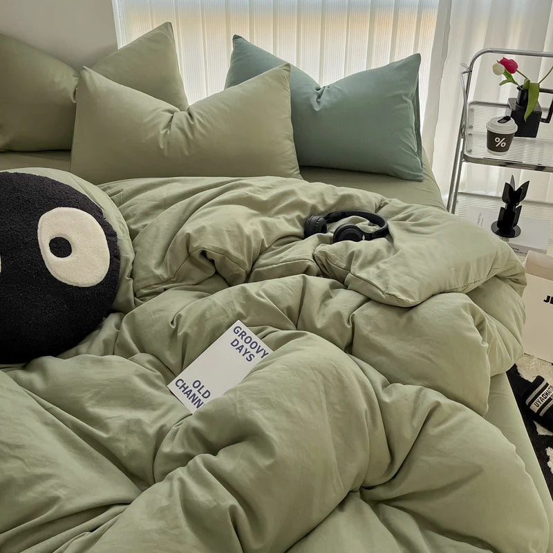 A bed with a duvet cover set in sage green and dusty blue tones, featuring pillows and a black stuffed animal.