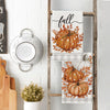 A set of three fabric kitchen towels featuring autumnal pumpkin and leaf designs on a wooden rack.