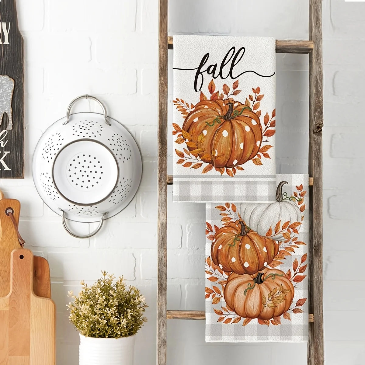 A set of three fabric kitchen towels featuring autumnal pumpkin and leaf designs on a wooden rack.