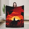 A person holds up a dark graphic blanket featuring a silhouette of a cowboy on a horse against a sunset background.