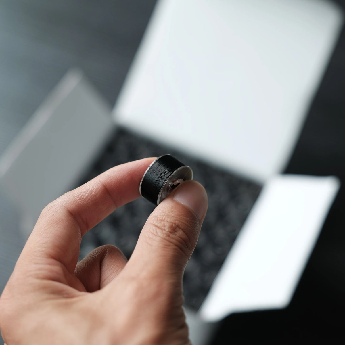 A hand holds a black embroidery bobbin thread against a blurred background of a laptop.