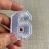 A hand holds a white embroidered number 6 patch for apparel.