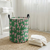 A green canvas laundry basket with pink bow patterns sitting on a woven mat next to a white shelf.