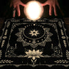 A hand holds a glowing orb above a dark mat decorated with celestial and floral tarot symbols.