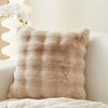 A fluffy beige faux fur throw pillow resting on a white surface with white flowers in the background.