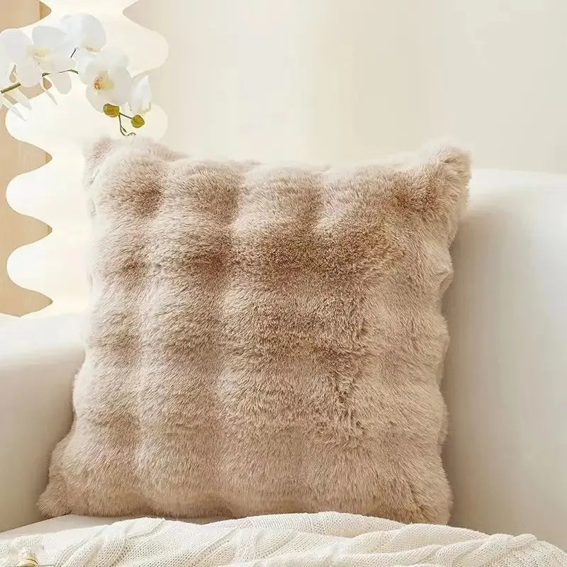 A fluffy beige faux fur throw pillow resting on a white surface with white flowers in the background.