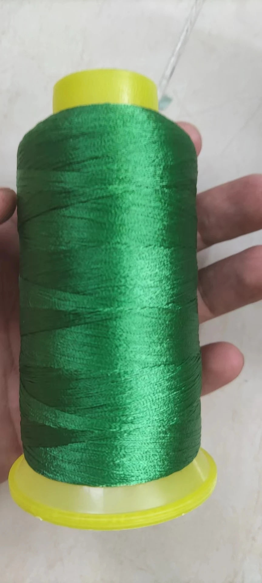 A hand holds a spool of bright green high tenacity polyester sewing thread with a yellow plastic base.