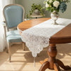 A round wooden dining table set with an embroidered white lace table runner and a light blue upholstered chair.