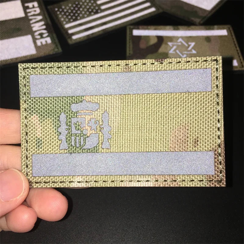 A hand holds a tactical multicam national flag patch with a crest and horizontal stripes.