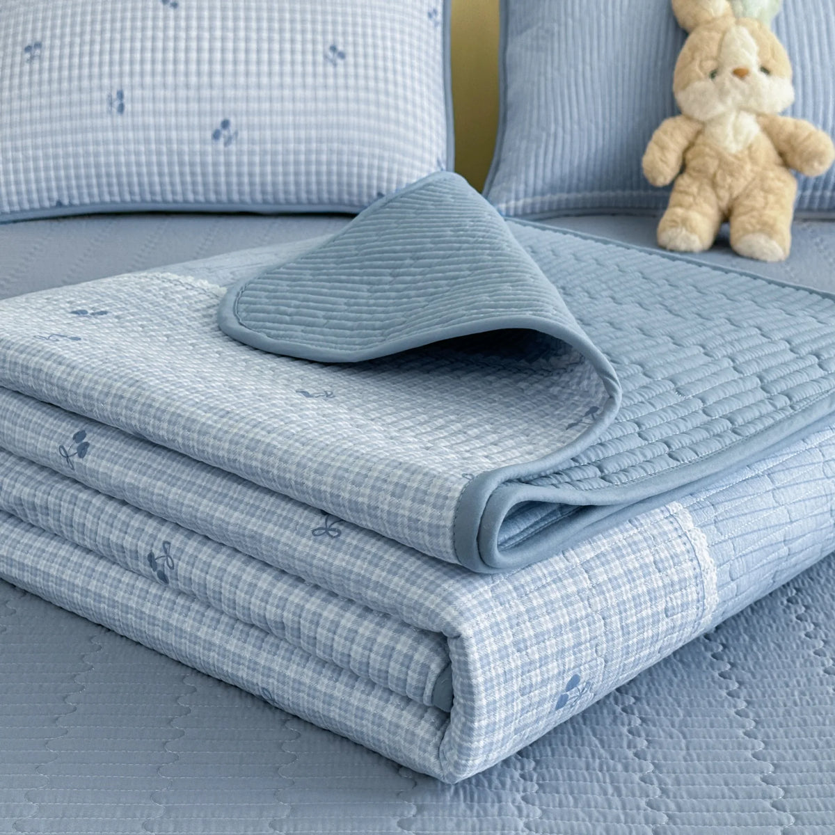 A light blue quilted bedspread with a subtle floral pattern is folded on a bed next to a small teddy bear.