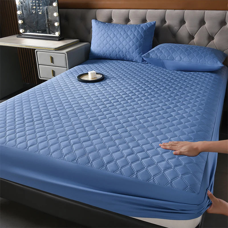 A blue quilted mattress protector is shown on a bed, with pillows and a bedside lamp visible.